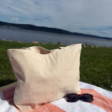 Lys canvas strandveske med Kreftforeningens logo, plassert på et håndkle i gresset ved vannkanten. Solbriller ligger ved siden av vesken.