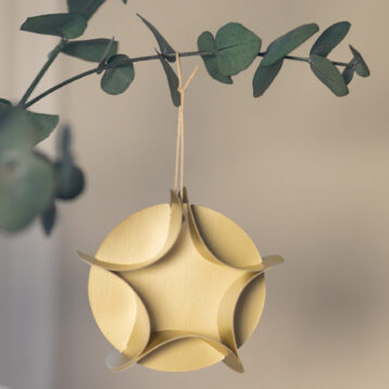 Et nærbilde av en dekorativ ornament laget av gullfarget papir, formet som en tredimensjonal stjerne med seks buede fliker som møtes i midten. Ornamentet henger i en tynn, lys snor festet til en gren med matte, grønne eucalyptusblader. Grenen strekker seg diagonalt fra venstre mot høyre, og bladene har en rund, flat form med en blågrønn tone. Bakgrunnen er mykt uskarp i beige og grå nyanser, noe som gir et minimalistisk og elegant uttrykk. Ornamentet har en matt overflate og fremstår som håndlaget, med en moderne og geometrisk design.