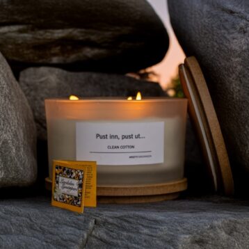 Et tent duftlys med etiketten "Pust inn, pust ut... CLEAN COTTON" står plassert mellom store grå steiner. Lokket ligger lent mot lyset, og foran står en liten gul eske med tekst og blomsterdekor. Stemningen er rolig og varm, med dempet kveldslys i bakgrunnen.